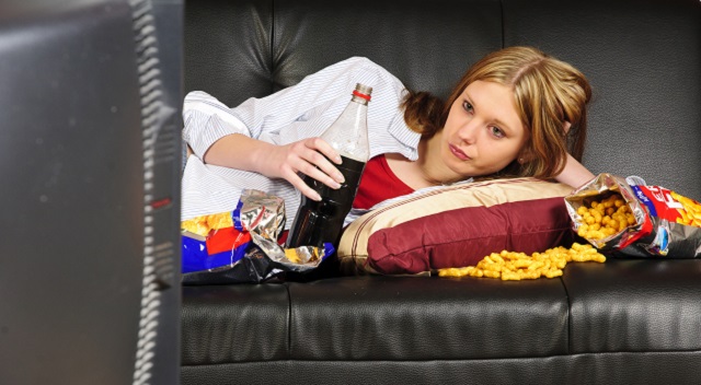 A young woman, teenager with long blond hair lolls on a black leather sofa, watching television and eating crisps and coke here.